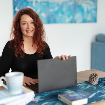 A woman with curly red hair smiles at a desk holding a laptop, books, and a cup in a well-lit room.