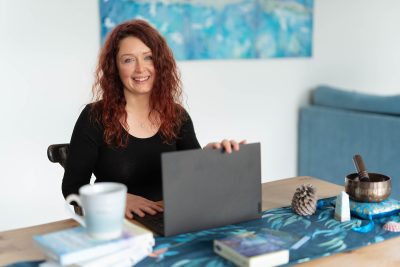 A woman with curly red hair smiles at a desk holding a laptop, books, and a cup in a well-lit room.