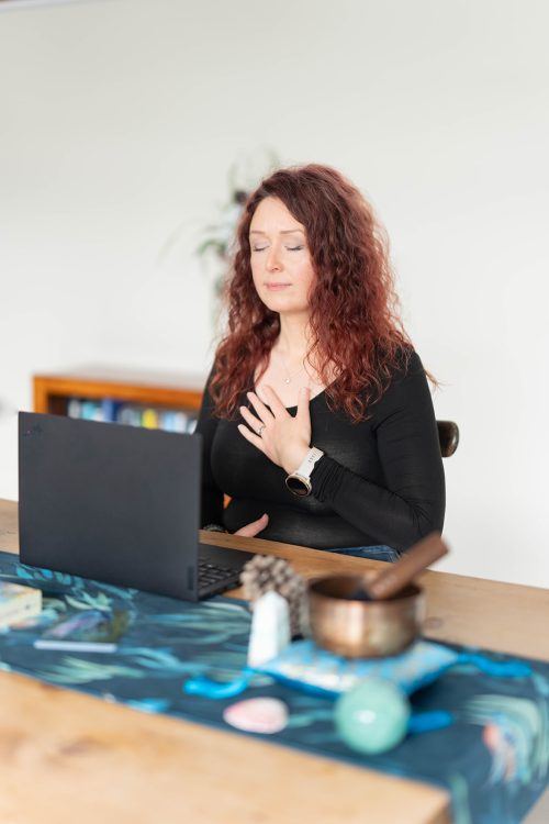 A woman with curly red hair sits at a table with her eyes closed, hand on chest, meditating in front of an open laptop.