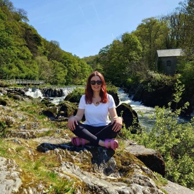 A woman with red hair sits cross-legged on rocks beside a river, with trees and a small building visible in the background.