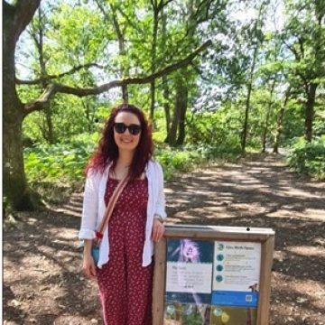 A woman wearing a red dress stands next to an informational sign along a sunlit woodland path bordered by trees.