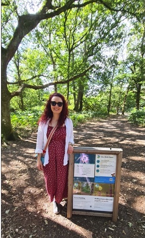 A woman wearing a red dress stands next to an informational sign along a sunlit woodland path bordered by trees.